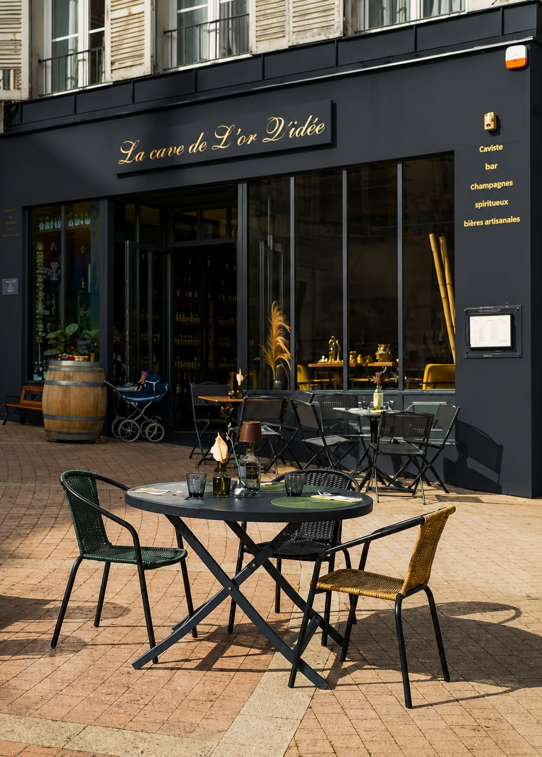 Devanture noire de La Cave de L'Or Q'idée, enseigne dorée et terrasse dressée sur le parvis au soleil