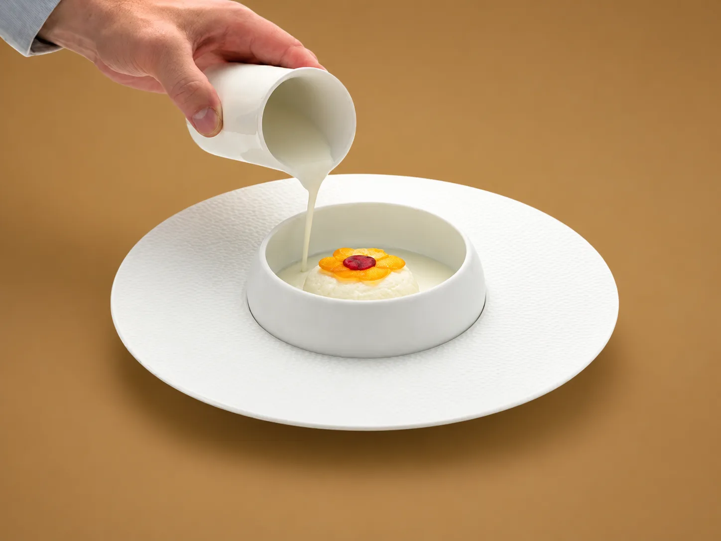 Dôme crémeux coiffé de pétales d'agrumes et d'un disque rouge, dans un bol blanc au centre d'une grande assiette de porcelaine grenue, pot de crème à côté